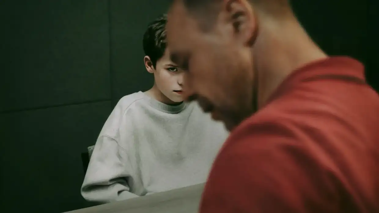 A serious-looking young boy in a grey sweatshirt sits at a table, staring intensely past a man in the foreground who is out of focus and dressed in red. The scene is dimly lit and suggests tension, possibly during an interrogation or difficult conversation.