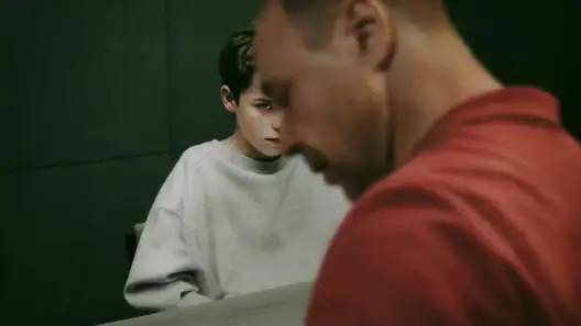 A serious-looking young boy in a grey sweatshirt sits at a table, staring intensely past a man in the foreground who is out of focus and dressed in red. The scene is dimly lit and suggests tension, possibly during an interrogation or difficult conversation.