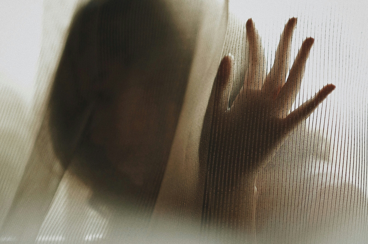 A blurred silhouette of a person stands behind a sheer, textured curtain. Their hand is pressed gently against the fabric, creating a sense of distance and obscurity. The lighting is soft and muted, giving the image a quiet, somber atmosphere.