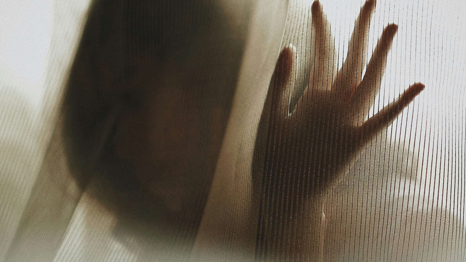 A blurred silhouette of a person stands behind a sheer, textured curtain. Their hand is pressed gently against the fabric, creating a sense of distance and obscurity. The lighting is soft and muted, giving the image a quiet, somber atmosphere.