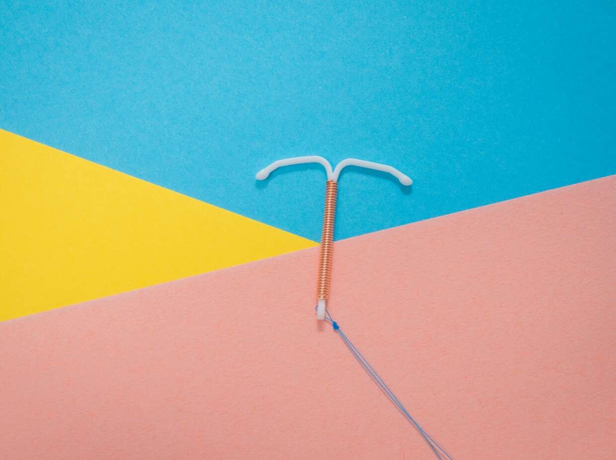 A copper intrauterine device (IUD) lies on a bright geometric background, with its blue removal strings trailing across the pink section.