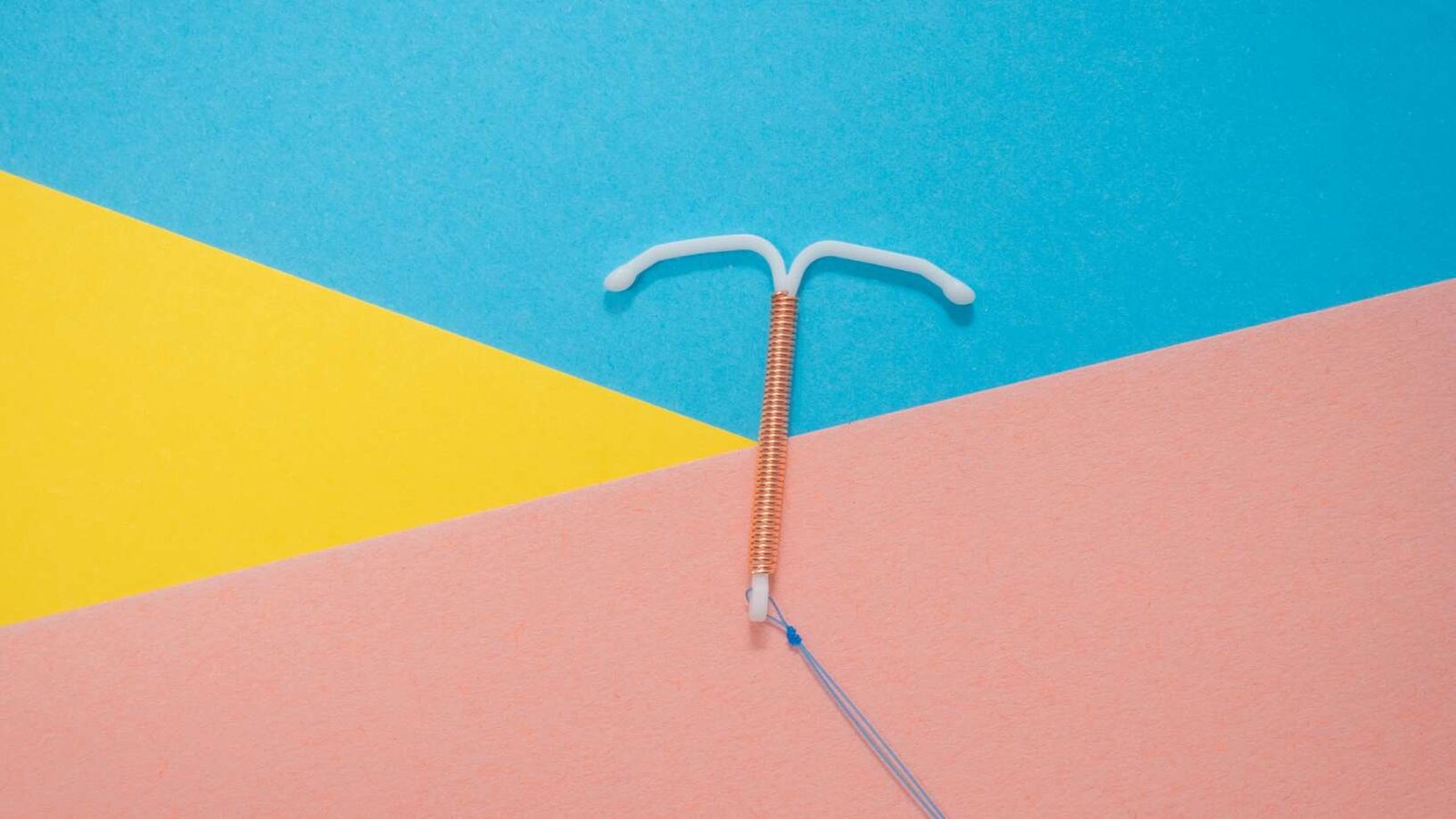 A copper intrauterine device (IUD) lies on a bright geometric background, with its blue removal strings trailing across the pink section.