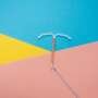 A copper intrauterine device (IUD) lies on a bright geometric background, with its blue removal strings trailing across the pink section.