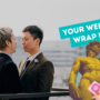 Two men stand closely facing each other on a rooftop, dressed in suits as if for a wedding or formal occasion, with a blurred cityscape in the background. On the right, an edited yellow-tinted image of Michelangelo’s David appears with a cartoon hand holding a pink condom in front of him. A bright teal banner above reads “YOUR WEEKLY WRAP UP.”