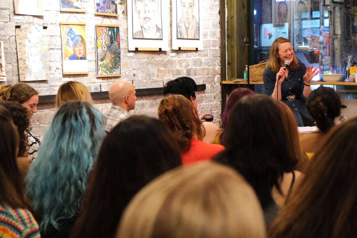 A person sits at the front of a crowded room, smiling and speaking into a microphone while gesturing with their hand. They face an audience seated closely together. The event takes place in a cosy indoor space with exposed brick walls and framed artwork hanging behind the speaker.