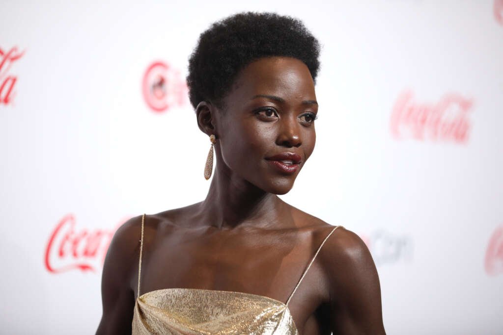 Lupita Nyong’o wearing a gold dress and drop earrings, posing on a red carpet against a branded backdrop.