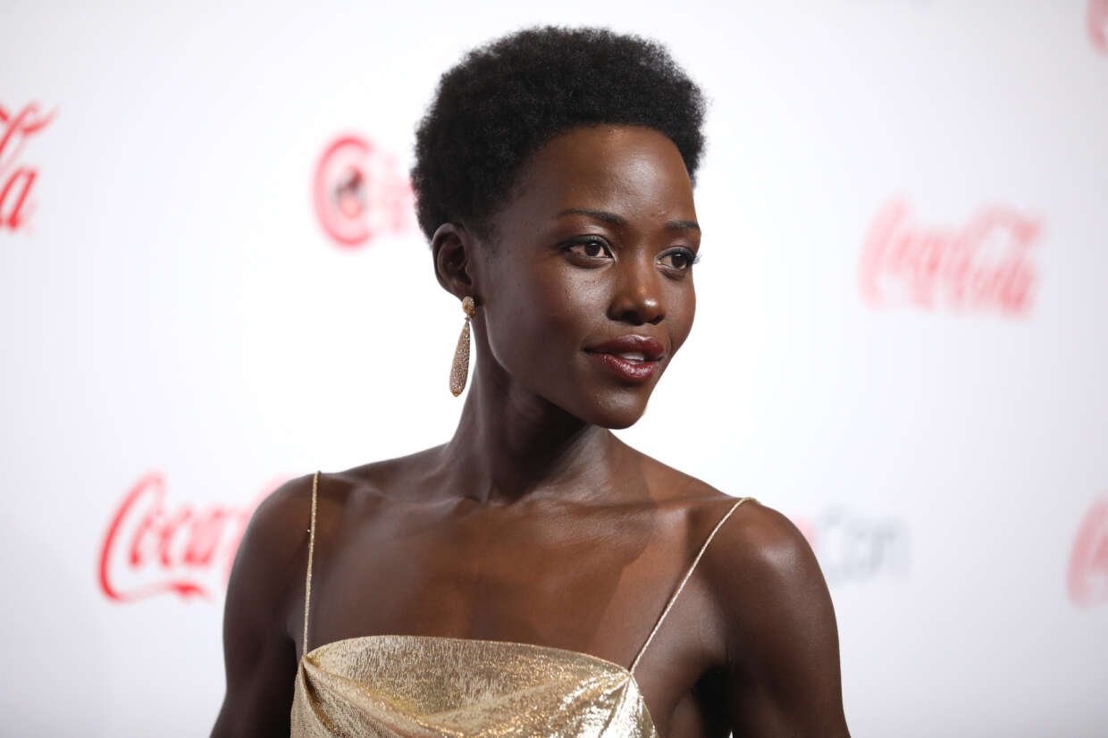 Lupita Nyong’o wearing a gold dress and drop earrings, posing on a red carpet against a branded backdrop.