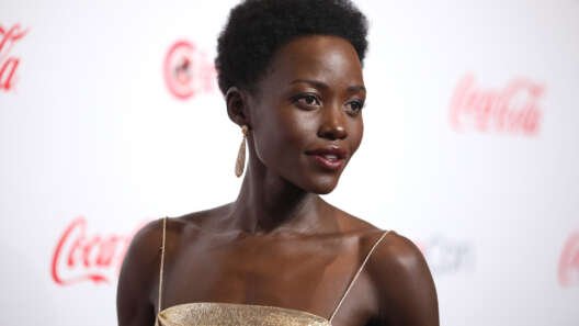 Lupita Nyong’o wearing a gold dress and drop earrings, posing on a red carpet against a branded backdrop.