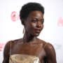 Lupita Nyong’o wearing a gold dress and drop earrings, posing on a red carpet against a branded backdrop.
