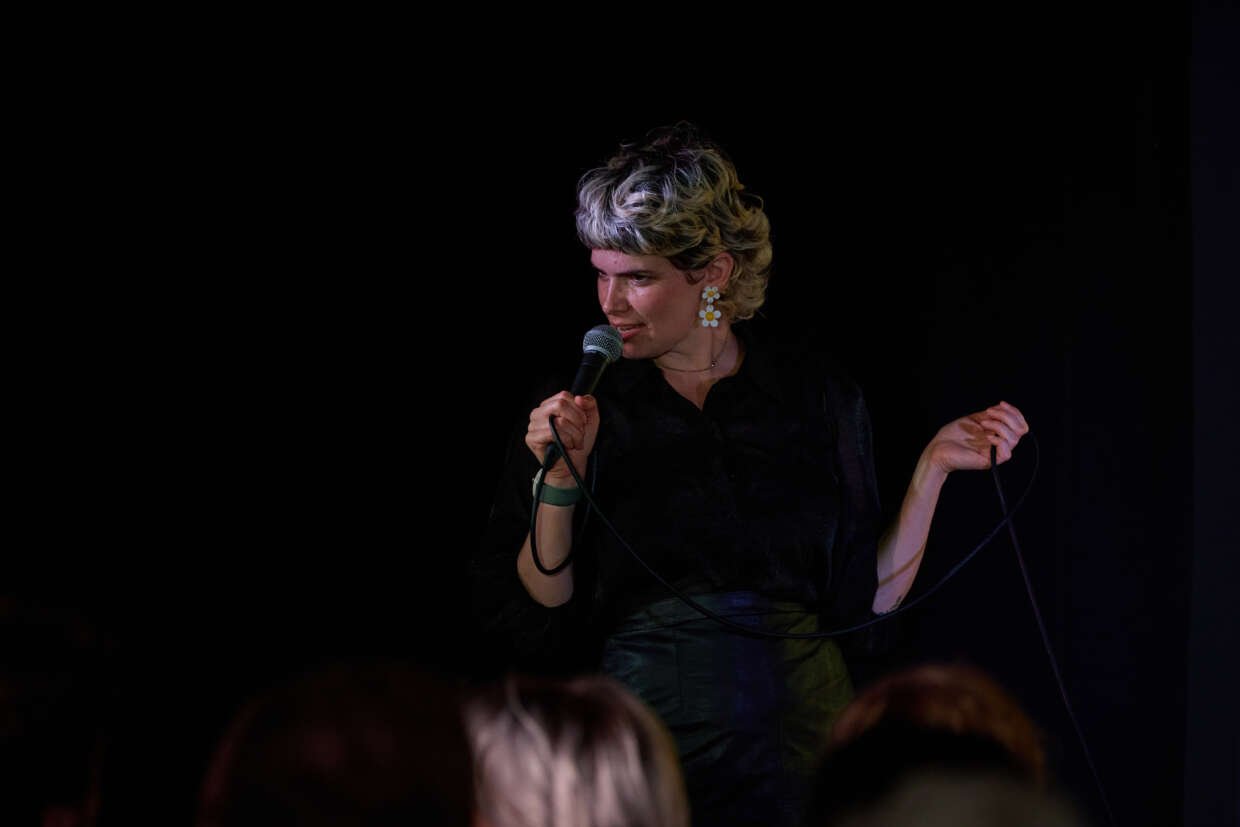Comedian with short light hair performing stand-up on stage, holding a microphone and cable in front of an audience.