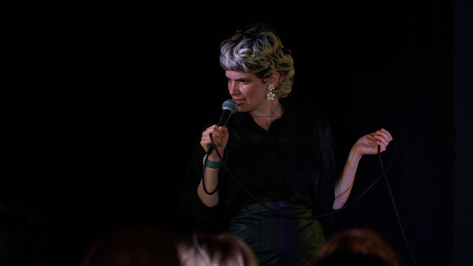 Comedian with short light hair performing stand-up on stage, holding a microphone and cable in front of an audience.
