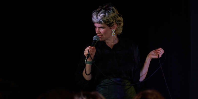 Comedian with short light hair performing stand-up on stage, holding a microphone and cable in front of an audience.