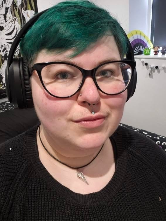 Person with short green hair wearing large round glasses and headphones, with a nose ring and lip piercing, sitting indoors with a decorative fan visible behind them.