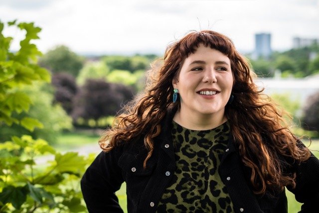 Person with long curly hair standing outdoors in a park, smiling with trees and greenery in the background.