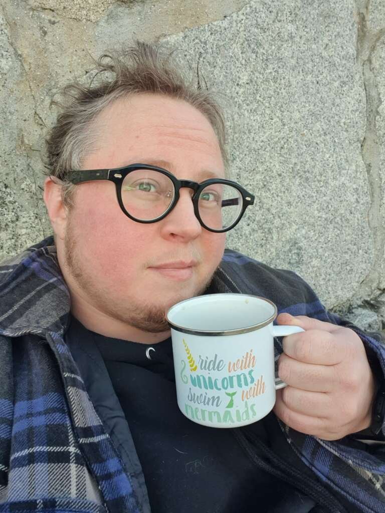 Person with short hair and round glasses holding a mug that reads “Ride with unicorns, swim with mermaids,” standing in front of a stone wall.