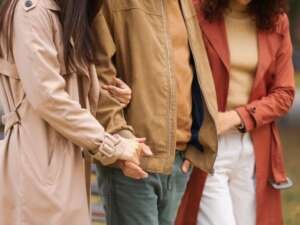 Three people walking outdoors side by side, arms linked, with two of them holding hands. They are wearing autumn coats in neutral tones.