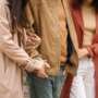 Three people walking outdoors side by side, arms linked, with two of them holding hands. They are wearing autumn coats in neutral tones.