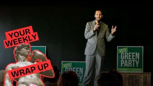 Man speaking on stage in front of “Vote Green Party” signs with audience in the foreground. Overlay graphic reads “Your Weekly Wrap Up” with a statue illustration and condom icon.