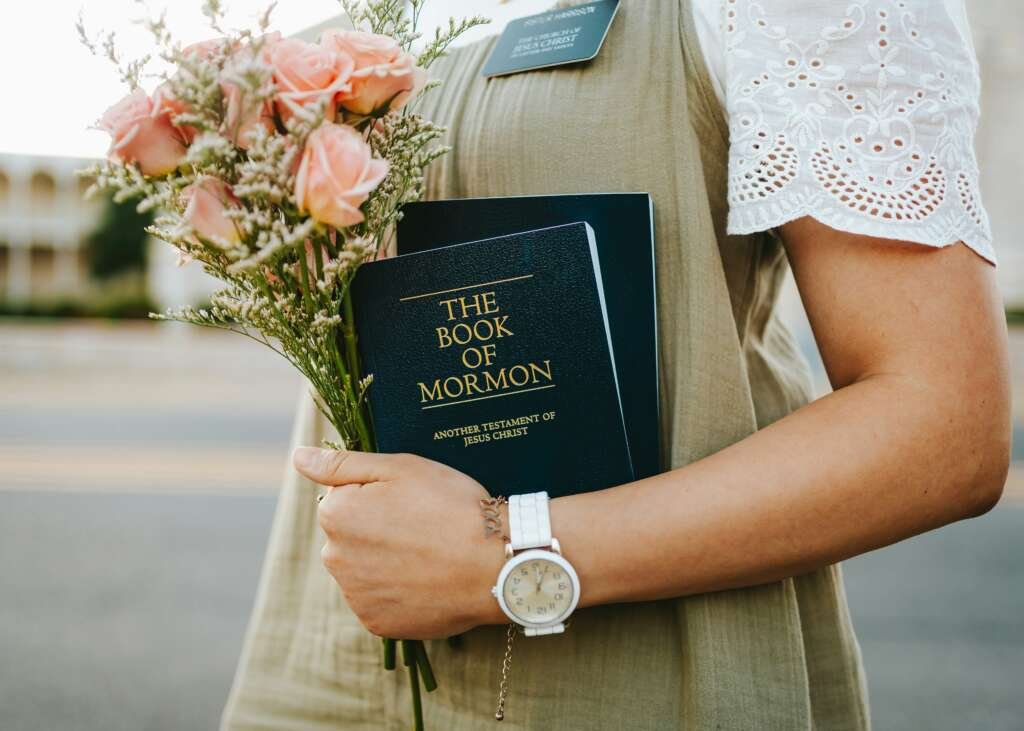 Person holding a bouquet of peach roses and a copy of The Book of Mormon, wearing a name badge and wristwatch outdoors.