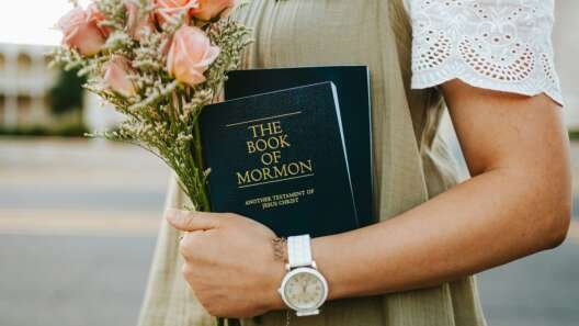 Person holding a bouquet of peach roses and a copy of The Book of Mormon, wearing a name badge and wristwatch outdoors.