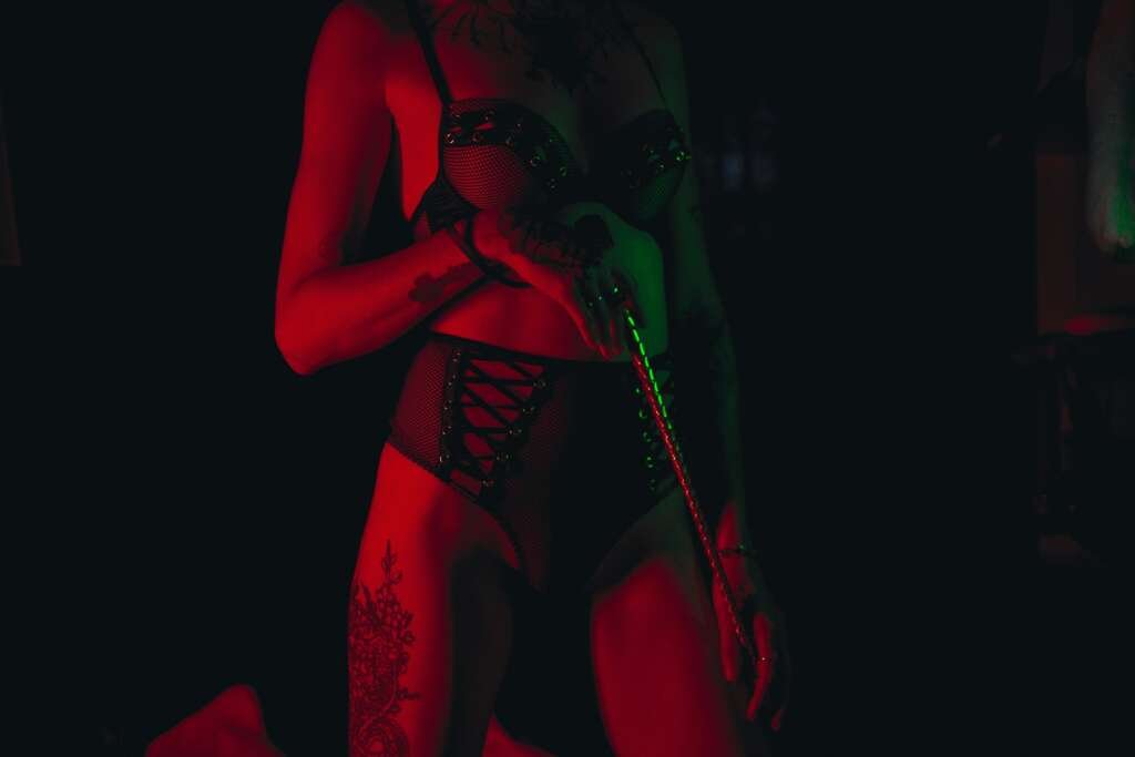Person wearing black lingerie under red and green lighting, holding a braided whip, with tattoos visible on their arm and thigh.