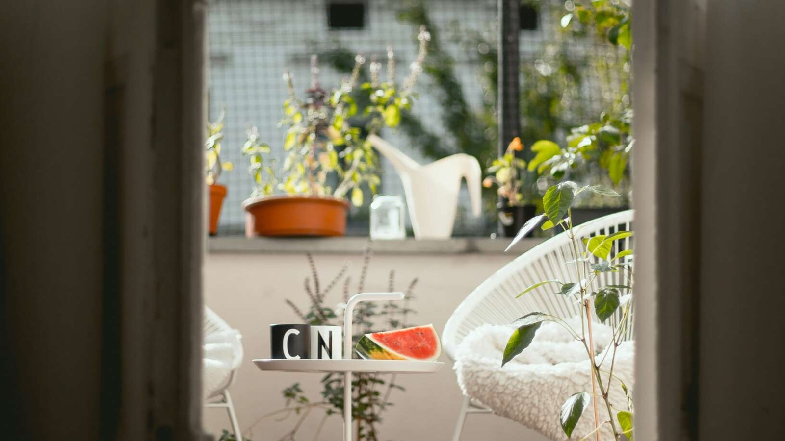 View through a doorway into a sunlit balcony with potted plants, a white chair with a cushion, and a small table holding a slice of watermelon and two mugs.