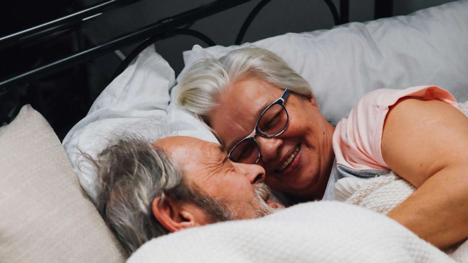 Older couple lying in bed together under a blanket, smiling and facing each other in a relaxed, affectionate moment.