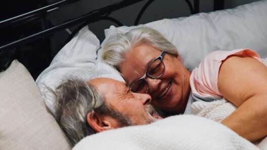 Older couple lying in bed together under a blanket, smiling and facing each other in a relaxed, affectionate moment.