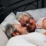 Older couple lying in bed together under a blanket, smiling and facing each other in a relaxed, affectionate moment.