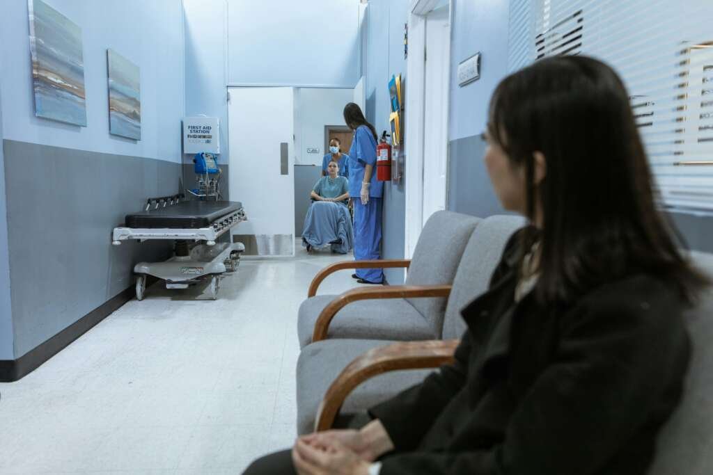 Hospital corridor with a woman seated in the foreground, looking toward a patient in a wheelchair being assisted by two healthcare workers in scrubs.