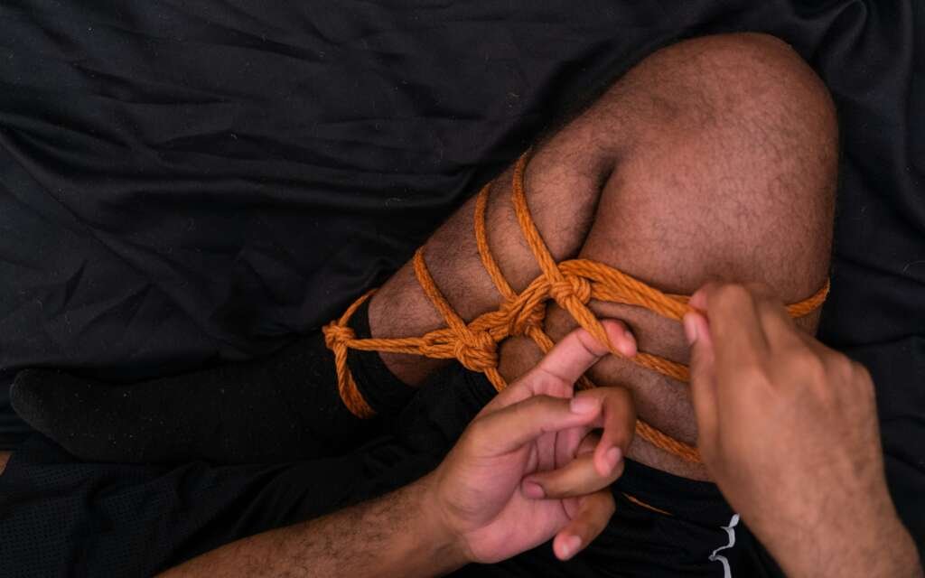 Close-up of hands tying orange rope around a person’s leg in a decorative knot against a dark fabric background.