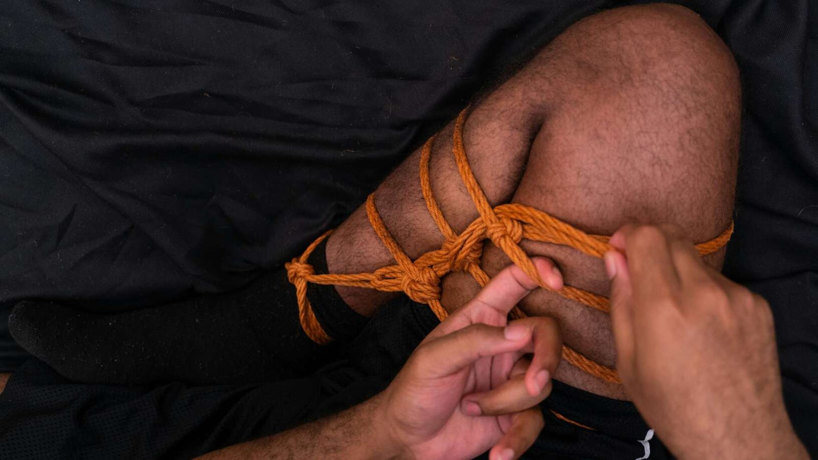 Close-up of hands tying orange rope around a person’s leg in a decorative knot against a dark fabric background.