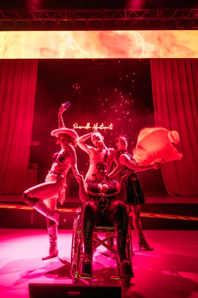 Group of performers posing on stage under red lighting, including a person in a wheelchair at the centre, wearing bold costumes and surrounded by theatrical props and bubbles.