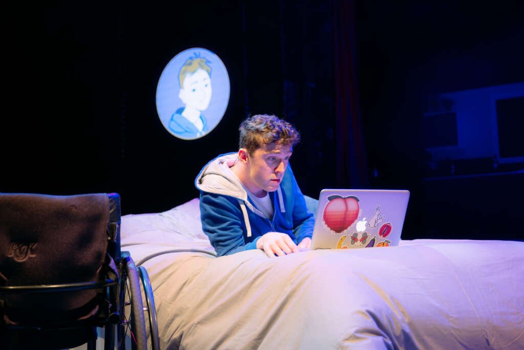 Young man lying on a bed using a laptop covered in stickers, with a wheelchair beside him and a projected cartoon avatar on the wall behind.
