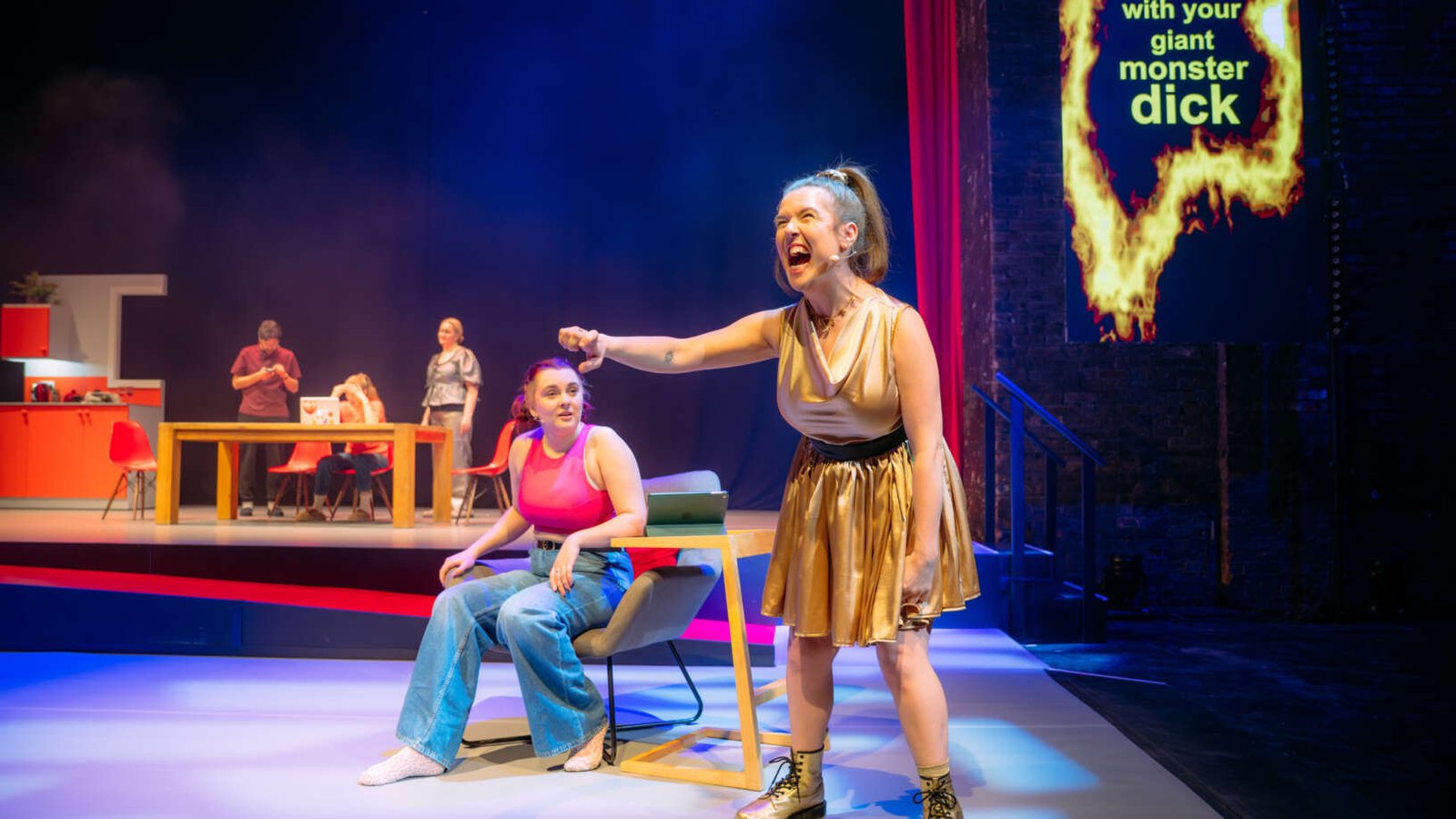 Theatre scene with a woman in a gold dress shouting and pointing while another woman sits beside her; other performers and a kitchen set are visible in the background, with bold projected text on the wall.