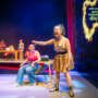Theatre scene with a woman in a gold dress shouting and pointing while another woman sits beside her; other performers and a kitchen set are visible in the background, with bold projected text on the wall.