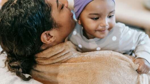 Adult holding a smiling baby wearing a purple headband and polka-dot outfit, wrapped together in a soft blanket indoors.