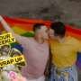 Two men kiss while holding a rainbow Pride flag behind them. A “Your Weekly Wrap Up” graphic featuring a classical statue and a condom illustration appears in the bottom left corner.