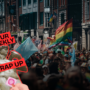 Crowds gather at a Pride march in a city street, with rainbow and transgender flags visible above the crowd. A “Your Weekly Wrap Up” graphic featuring a classical statue and a condom illustration appears in the bottom left corner.