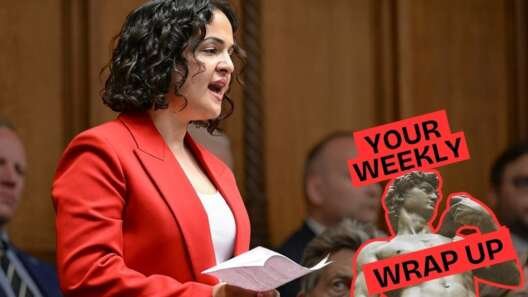 A person wearing a bright red blazer speaks while holding papers in a parliamentary chamber, with seated people visible in the background. A “Your Weekly Wrap Up” graphic featuring a classical statue and a condom illustration appears in the bottom right corner.