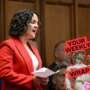 A person wearing a bright red blazer speaks while holding papers in a parliamentary chamber, with seated people visible in the background. A “Your Weekly Wrap Up” graphic featuring a classical statue and a condom illustration appears in the bottom right corner.