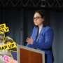 A promotional graphic for Pillow Talk Scotland's "Your Weekly Wrap Up." The background shows a woman in a bright blue blazer speaking at a lectern with microphones, gesturing as she addresses an audience. In the bottom left corner is the recurring "Your Weekly Wrap Up" branding featuring Michelangelo's David statue holding a condom packet, with yellow labels reading "YOUR WEEKLY" and "WRAP UP."