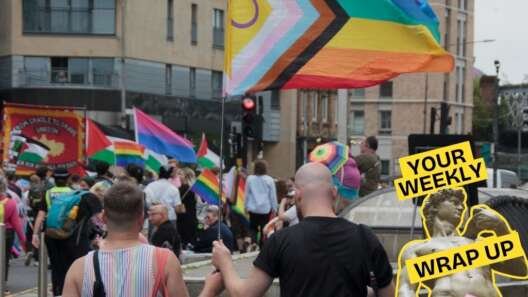 A crowd marches through Glasgow during a Pride event, with people carrying rainbow, Progress Pride, bisexual and Palestinian flags. In the foreground, two people walk hand in hand beneath a large Progress Pride flag. Overlaid on the image is Pillow Talk Scotland's "Your Weekly Wrap Up" graphic featuring Michelangelo's David statue, bold yellow text and a pink condom illustration.