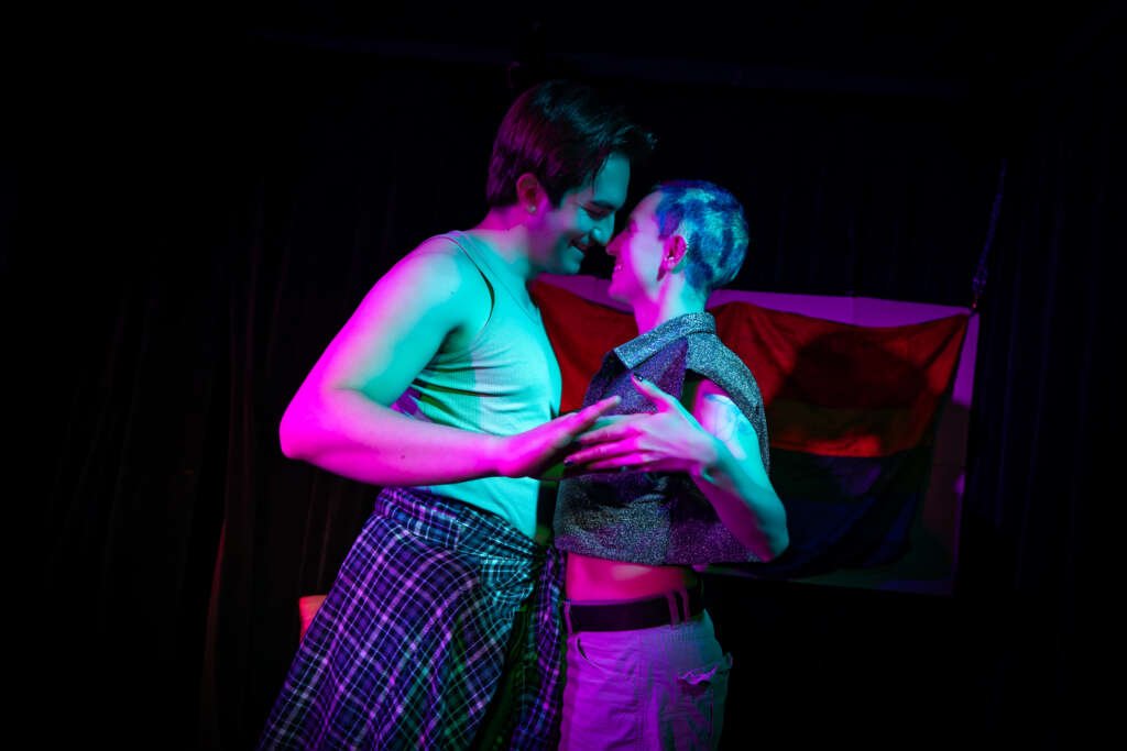Two performers stand close together under vivid pink and blue stage lighting, smiling at each other with their faces almost touching. A rainbow flag hangs behind them.