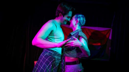 Two performers stand close together under vivid pink and blue stage lighting, smiling at each other with their faces almost touching. A rainbow flag hangs behind them.