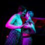 Two performers stand close together under vivid pink and blue stage lighting, smiling at each other with their faces almost touching. A rainbow flag hangs behind them.