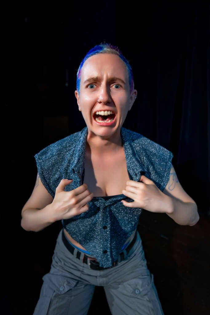 A performer with very short hair, a nose ring and visible arm tattoos grips a sparkly sleeveless top and leans towards the camera with an exaggerated open-mouthed expression against a dark background.