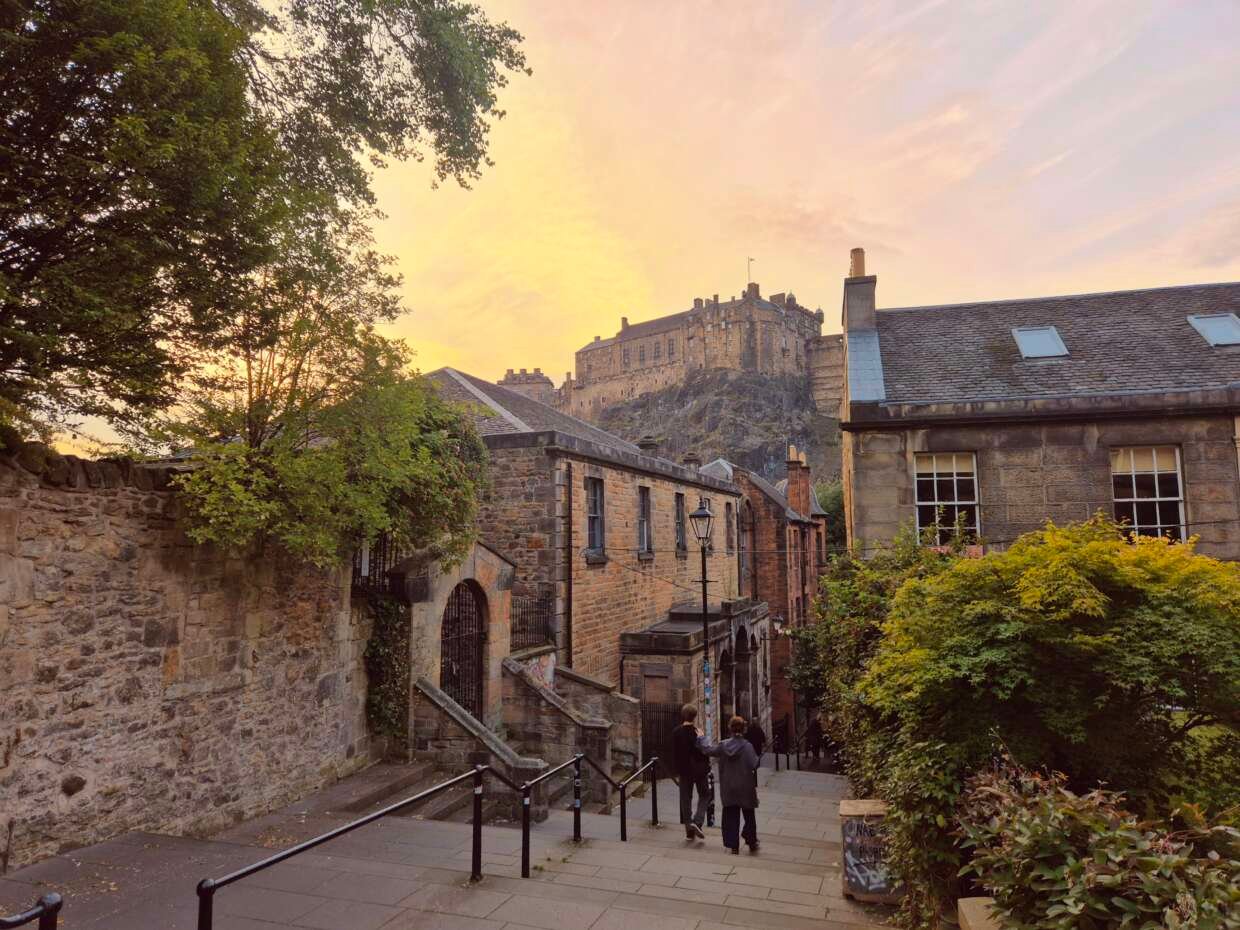 Edinburgh Castle seen at sunset from a steep Old Town street, framed by historic stone buildings, leafy trees and greenery. Two people walk downhill in the foreground beneath a warm orange and pink sky.