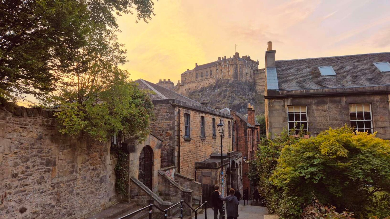 Edinburgh Castle seen at sunset from a steep Old Town street, framed by historic stone buildings, leafy trees and greenery. Two people walk downhill in the foreground beneath a warm orange and pink sky.