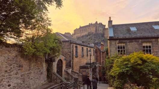 Edinburgh Castle seen at sunset from a steep Old Town street, framed by historic stone buildings, leafy trees and greenery. Two people walk downhill in the foreground beneath a warm orange and pink sky.